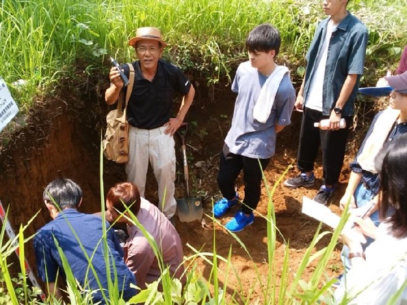 つくば台地の黒ボク土断面に触れながら土壌の成り立ちの解説を受ける学生たち。