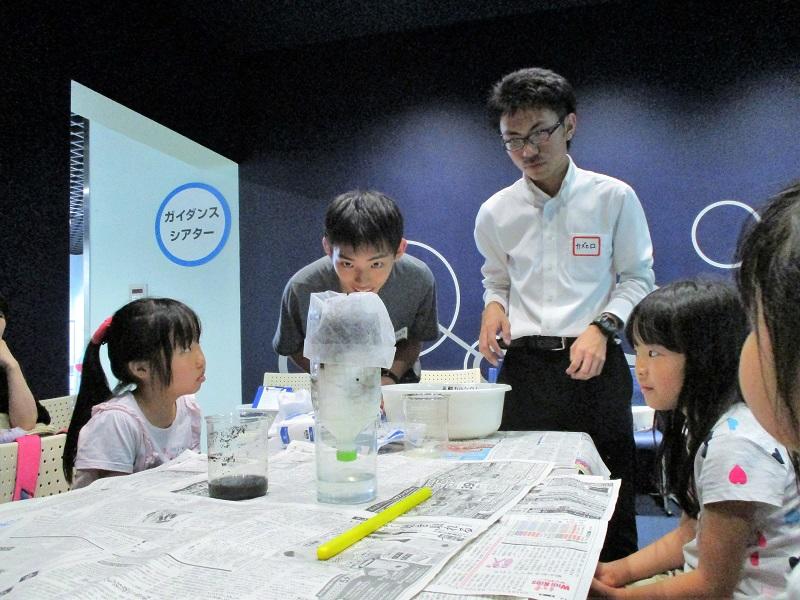 「水の汚れはどこへ消える？ (浄化)」という実験で炭と脱脂綿を用いて汚れた水がきれいになるのか見守る高校生(写真奥左 )、大学生(写真奥右)と小学生ら。