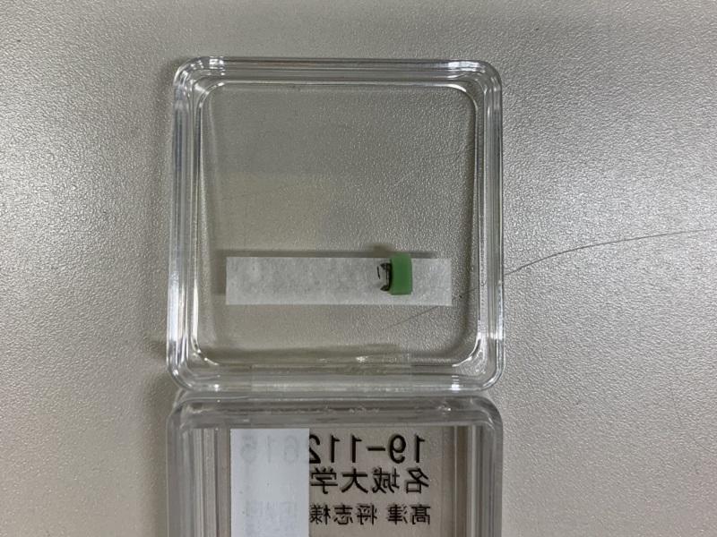 MSTのFIB装置によって薄片化加工された AlInN/GaN多層膜反射鏡試料であり、試料は縦10µm、横20µm、厚さ80～90nm程度である。Mo製のメッシュに貼り付けられたカーボンの土台に固定されている。大きな振動を与えると試料が壊れてしまうため、気を付けて運んだ。