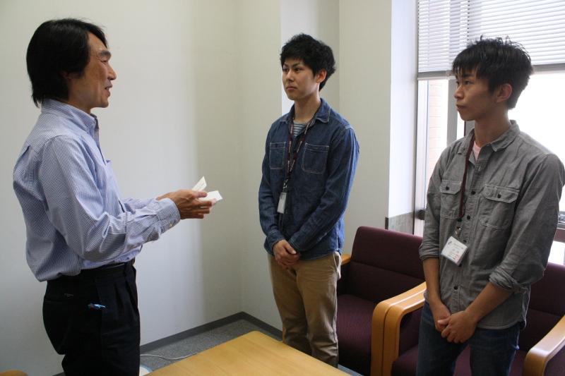 勝浦部長から義援金を受け取る岩田さん（右）と高井さん（学務センターで）