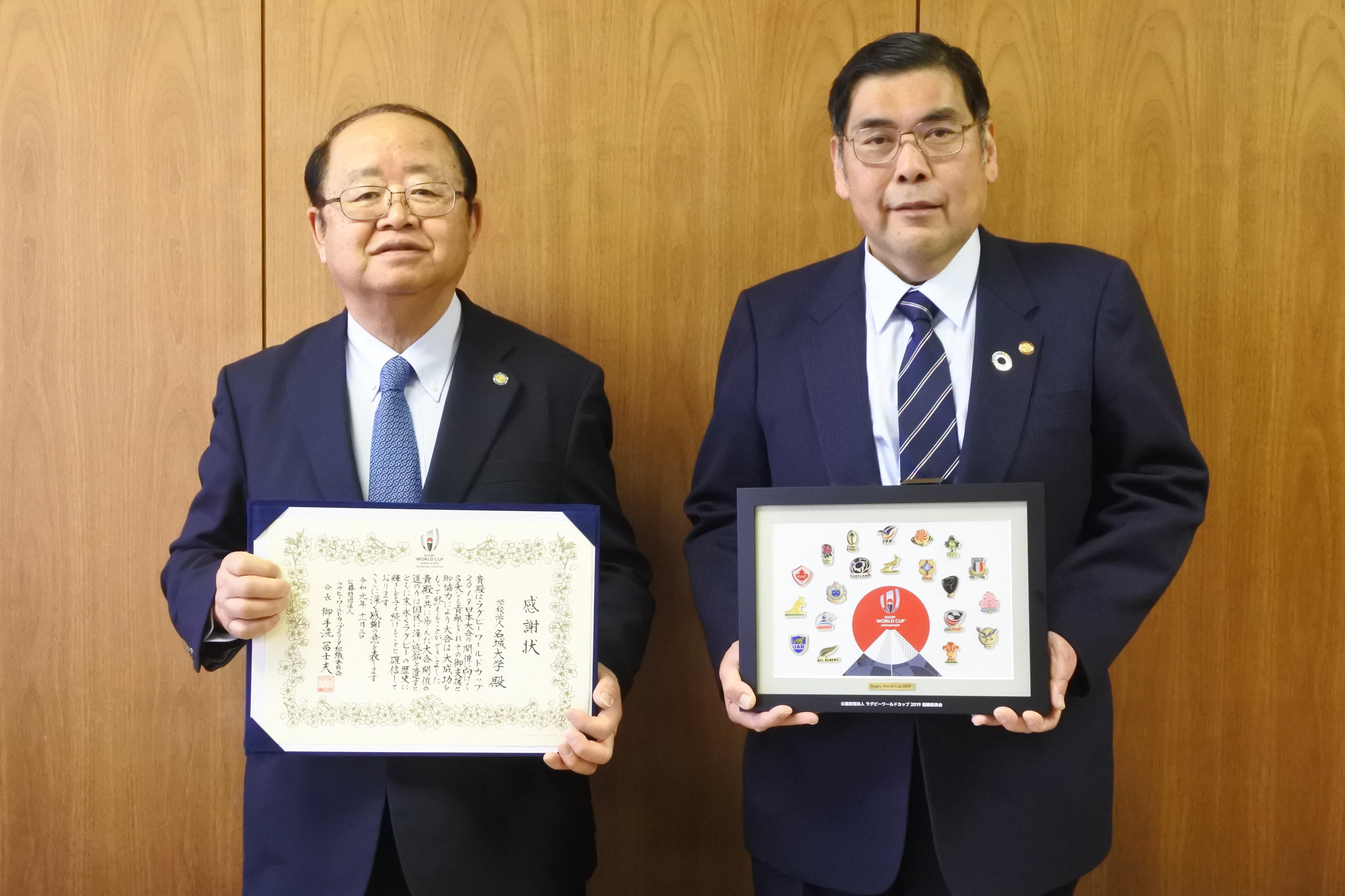 感謝状と記念のピンバッジ額を持つ立花貞司理事長（左）と小原章裕学長