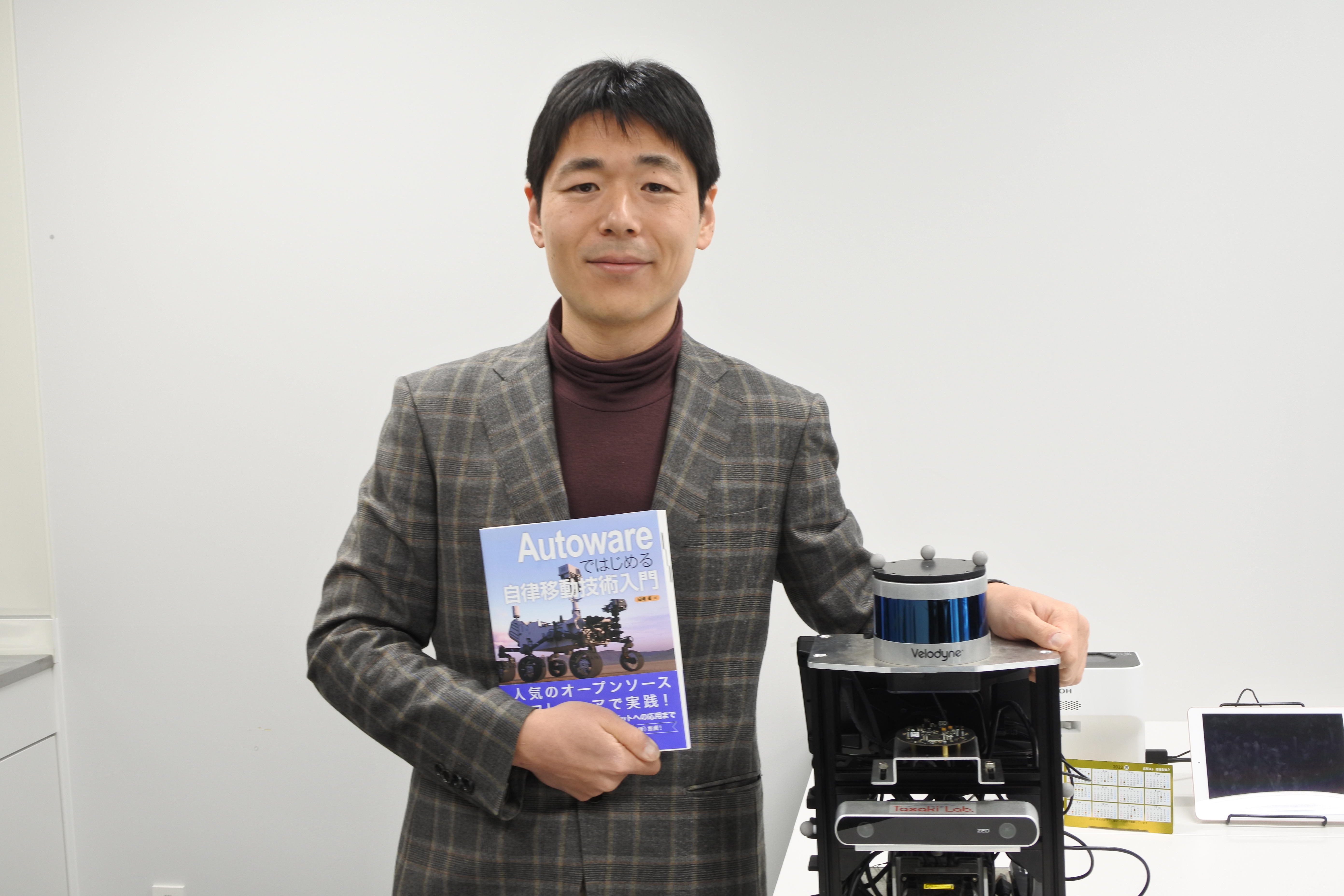 天白キャンパスの研究室にあるロボットと一緒に写真に納まる田崎豪准教授